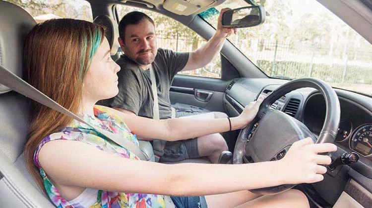 一个十几岁的女孩坐在汽车的驾驶座上，双手放在方向盘上. 她父亲坐在副驾驶座位上调整后视镜. 